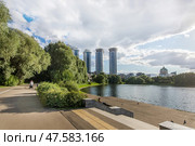 People stroll along a winding path near a serene lake surrounded by lush greenery. Tall modern buildings rise in the background under a partly cloudy sky. (2018 год). Стоковое фото, фотограф Алексей Климкин / Фотобанк Лори
