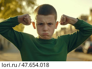 Kid boy shows his muscles. Angry little child boy waves his fist at the camera outdoors at the city street. Unhappy grumpy boy showing his biceps. Provocation. Elementary school problems. Bullying. Стоковое AI-изображение, AI-художник Марина Демидюк / Фотобанк Лори