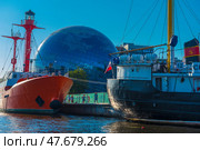 A striking and recognizable scene in Kaliningrad is the museum complex on the embankment. In the foreground are two ships: a red lighthouse ship and a black and white fishing vessel "CPT-129". In the center of the frame, against the backdrop of the ships, rises a spherical building - this is the Museum of the World Ocean with its famous dome-planetarium, lined with mirrored glass. Around - modern urban architecture, water and a clear blue sky. (2020 год). Редакционное фото, фотограф Анна Кривицкая / Фотобанк Лори