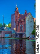 A picturesque red brick Gothic building standing right on the river bank in Kaliningrad. The corner turret with a high spire and decorative battlements give the building the appearance of a small castle or watchtower. The building can be recognized as one of the old water towers or security structures typical of Königsberg architecture. In the foreground is a water surface with a reflection of the building, and on the right are people relaxing with a dog. In the background are modern and historical brick houses. Стоковое фото, фотограф Анна Кривицкая / Фотобанк Лори