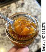 A close-up of a jar of amber-colored homemade jam or marmalade, shot in natural light. In the foreground is a spoon filled with thick, aromatic mass with pieces of fruit, located directly above the jar. The texture of the jam is delicious, shiny and viscous, giving the image warmth and coziness. In the background is a blurred stone countertop. An ideal shot for food blogs, recipes, gastronomic projects, advertising of home-cooked food and farm products. Стоковое AI-изображение, AI-художник Анна Кривицкая / Фотобанк Лори