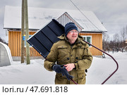 Carrying wide snow shovel over shoulder mature white man in warm coat and knitted hat appears focused while standing near wooden house in snowy setting. Стоковое фото, фотограф Владимир Григорьев / Фотобанк Лори