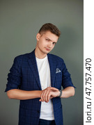 A young man standing confidently, adjusting his watch against a neutral background. He is dressed in a fashionable blue blazer and white shirt, exuding a sense of professionalism and style in Dubai. Стоковое фото, фотограф Aleksandr Golubev / Фотобанк Лори