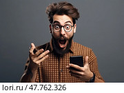 Startlingly surprised dapper young man with beard and mouth open looks away holding phone against gray background. Стоковое AI-изображение, AI-художник Данила Большаков / Фотобанк Лори
