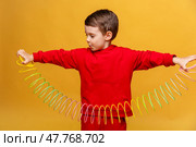 Happy smiling little boy 3-4 years old, wearing fashion red sport suit, plays with a plastic colored neon rainbow spiral children toy, holding bouncy stretchy toy on yellow background, copy space. Стоковое фото, фотограф Ирина Михайличенко / Фотобанк Лори