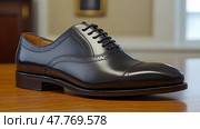 A polished black dress shoe rests on a wooden table, showcasing its sleek design and reflecting warm sunlight in an elegant indoor space. Стоковое AI-изображение, AI-художник Данила Большаков / Фотобанк Лори