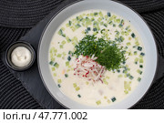 Cold Russian soup okroshka featuring tangy kefir base, diced cucumbers, radishes, fresh herbs, topped with creamy sour cream garnish. Стоковое фото, фотограф Данил Руденко / Фотобанк Лори
