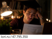 Graduate studious in dimly lit room reflects on coursework with focused expression and graduation cap. Стоковое AI-изображение, AI-художник Сергей Толмачёв / Фотобанк Лори
