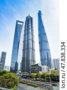 Shanghai, China - 1 April 2025: Lujiazui's iconic skyline: Shanghai Tower, Jin Mao Tower, and Shanghai World Financial Center, framed by greenery and urban infrastructure under blue skies. Редакционное фото, фотограф Татьяна Терехина / Фотобанк Лори
