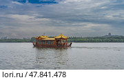 A beautiful Chinese wooden boat floats on the lake. Curved roof edges, latticed railings, silhouettes of people on the deck. Green vegetation on the shore. Blue sky, clouds. China. Hangzhou. West Lake. Стоковое фото, фотограф Вера Ивановна Кубарева / Фотобанк Лори