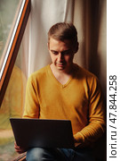 Bathed in warm sunlight, a young man sits comfortably by the window, focused on his laptop. The tranquil setting is enhanced by soft curtains and a relaxed atmosphere, perfect for creativity. Стоковое фото, фотограф Aleksandr Golubev / Фотобанк Лори
