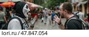 A street performer in a striking mask engages in a lively conversation with a captivated onlooker in an urban setting. Стоковое AI-изображение, AI-художник Данила Большаков / Фотобанк Лори
