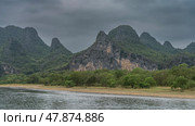 Picturesque mountains against the sky and clouds. Green vegetation on steep slopes and on the river bank. The riverbed is in the foreground. China. Li River. Li Jiang. Guilin. Стоковое фото, фотограф Вера Ивановна Кубарева / Фотобанк Лори