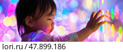 A child with dark hair touches a brightly lit wall, mesmerized by the colorful lights around them in a playful environment. Стоковое AI-изображение, AI-художник Данила Большаков / Фотобанк Лори