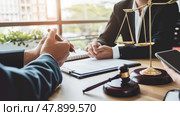 Two individuals are seated at a table located in front of a scale of justice, engaging in discussions or negotiations. The symbolic representation of justice is prominent in the background. Стоковое AI-изображение, AI-художник Данила Большаков / Фотобанк Лори