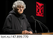 Elderly woman standing confidently at a podium indoors, addressing an unseen audience through a microphone. Стоковое AI-изображение, AI-художник Данила Большаков / Фотобанк Лори
