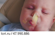 A baby with food stained face trying to eat with a spoon while sitting in a highchair. Close-up. A blue-eyed baby eats porridge with a spoon. Useful food. Стоковое видео, видеограф Воловодов Владимир / Фотобанк Лори