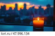 A candle in a glass burns brightly near a window with a city skyline at dusk during a power outage. Стоковое AI-изображение, AI-художник Igor Tichonow / Фотобанк Лори