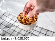 Hand picking an almond from a white ceramic bowl overflowing with almonds, resting on a checkered tablecloth atop a light wooden table, highlighting healthy snacking options. Стоковое фото, фотограф Ольга Ильина / Фотобанк Лори