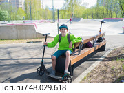 12-летний мальчик с травмированными коленями сидит на скамейке с самокатом, на фоне памп-трека для катания на скейтбордах и самокатах. Подросток в экстрим кластере на Лукинской улице в Москве. Стоковое фото, фотограф Яна Проничева / Фотобанк Лори