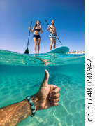 Split-shot photograph taken below the water surface. Thumbs up hand is underwater, two happy people on boards over water. Стоковое AI-изображение, AI-художник Катерина Ковалева / Фотобанк Лори
