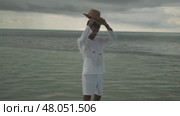 A young man in light white clothes and a hat walks barefoot in the rain on the water of the sea. Stormy evening clouds are in the background. Maldives. Стоковое видео, видеограф Aleksey Kidisyuk / Фотобанк Лори