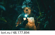 A child cautiously explores a dark jungle, holding a flashlight and studying a map. Стоковое AI-изображение, AI-художник Данила Большаков / Фотобанк Лори