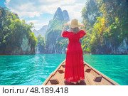 Woman female traveler enjoys the breathtaking views of Cheow Lan Lake, surrounded by towering limestone cliffs and lush greenery in Khao Sok, Thailand. Wearing a vibrant red dress and wide-brimmed hat. Стоковое фото, фотограф Владимир Разгуляев / Фотобанк Лори