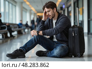 Мужчина сидит на полу рядом с чемоданом, явно уставший от долгого ожидания или перелета. Стоковое AI-изображение, AI-художник Валерий Лукин / Фотобанк Лори