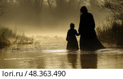 A woman holds a child's hand as they wade through a serene wetland, enveloped in morning mist and illuminated by soft light. Стоковое AI-изображение, AI-художник Данила Большаков / Фотобанк Лори