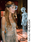 A young woman engages with an augmented reality display in a tech exhibit, captivated by holographic figures and digital innovations. Стоковое AI-изображение, AI-художник Данила Большаков / Фотобанк Лори