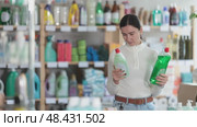 Female customer stands in the household section of a supermarket and chooses between a regular and eco-friendly dishwashing detergent. Woman shopping for household products for the home. Стоковое видео, видеограф Яков Филимонов / Фотобанк Лори