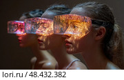Three women engage with a captivating art piece, their visors reflecting vibrant colors in a dim gallery during an evening event. Стоковое AI-изображение, AI-художник Данила Большаков / Фотобанк Лори
