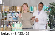 Attentive young woman scrolling phone while looking for something and pharmacist consulting her in drugstore. Стоковое видео, видеограф Яков Филимонов / Фотобанк Лори