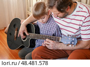 Handsome dad teaches his cute son to play the guitar sitting on sofa in the living room at home. Happy Father's Day. Father teaching his child to play guitar. Kid Learning music skill. Art education. Стоковое фото, фотограф Марина Демидюк / Фотобанк Лори