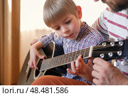 Handsome dad teaches his cute son to play the guitar sitting on sofa in the living room at home. Happy Father's Day. Father teaching his child to play guitar. Kid Learning music skill. Art education. Стоковое фото, фотограф Марина Демидюк / Фотобанк Лори