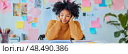 Creative burnout: Young woman with curly afro, struggling with idea block, rests her head on her hands at a cluttered desk in a vibrant office. Стоковое AI-изображение, AI-художник Данила Большаков / Фотобанк Лори