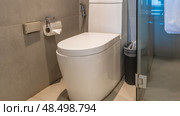 A toilet in the corner of the bathroom. The lid is closed, lowered.The urn is nearby. There is a roll of toilet paper on the beige tiled wall. The shower cabin is behind glass partitions.A white towel. Стоковое фото, фотограф Вера Ивановна Кубарева / Фотобанк Лори