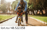 A father is guiding his young son as he learns to ride a bike for the first time. The father holds onto the bikes handlebars, assisting the boy in finding his balance. Стоковое AI-изображение, AI-художник Данила Большаков / Фотобанк Лори