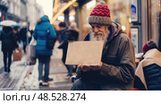 An elderly man sits on a busy street corner in the evening, holding a blank cardboard sign, as people pass by him. Стоковое AI-изображение, AI-художник Данила Большаков / Фотобанк Лори