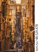 VALLETTA, MALTA - NOVEMBER 12, 2018 - Hilly ancient streets view with colourful balconies, people and cars in the medieval city. Редакционное фото, фотограф Сергей Богомяко / Фотобанк Лори