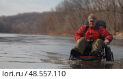 An elderly gentleman propels his adapted wheelchair through shallow waters, surrounded by a serene autumnal backdrop. Стоковое AI-изображение, AI-художник Данила Большаков / Фотобанк Лори