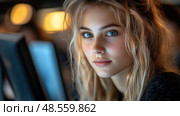 A young woman with beautiful blue eyes looks towards the viewer, illuminated by soft light from nearby screens in a darkened setting. Стоковое AI-изображение, AI-художник Данила Большаков / Фотобанк Лори