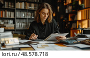 A woman focuses intently on financial documents, calculating figures at her cluttered desk filled with papers and surrounded by shelves of books. Стоковое AI-изображение, AI-художник Данила Большаков / Фотобанк Лори