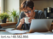 Усталый фрилансер  с чашкой кофе,  работая за ноутбуком в уютном домашнем офисе. Стоковое AI-изображение, AI-художник Елена Болотина / Фотобанк Лори