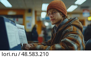 A young adult attentively completes paperwork while seated at a desk, surrounded by a bustling indoor setting filled with people. Стоковое AI-изображение, AI-художник Данила Большаков / Фотобанк Лори