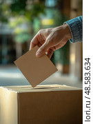 A hand inserts a ballot into a cardboard box in a polling station, highlighting civic engagement during the local election. Стоковое AI-изображение, AI-художник Данила Большаков / Фотобанк Лори
