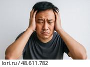 Asian man clutching head with both hands eyes closed expressing deep pain frustration tension against plain white background. Стоковое AI-изображение, AI-художник Павел Муравьев / Фотобанк Лори