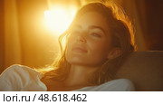 A young woman relaxes, soaking in the warm glow of the sunlight filling her cozy room at sunset, embodying tranquility and contentment. Стоковое AI-изображение, AI-художник Данила Большаков / Фотобанк Лори