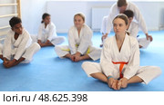 Group of positive tweens of different nationalities wearing white kimonos sitting on tatami in gym, performing butterfly stretch during martial arts workout with coach. Стоковое видео, видеограф Яков Филимонов / Фотобанк Лори